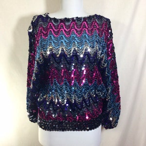 May include: A sequined top with a wavy pattern in shades of pink, blue, and black. The top has long sleeves and a scoop neck.