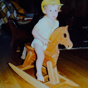 May include: A young child wearing a yellow and green baseball cap rides a wooden rocking horse. The horse is brown and has a white saddle. The child is wearing a white shirt with a yellow collar.