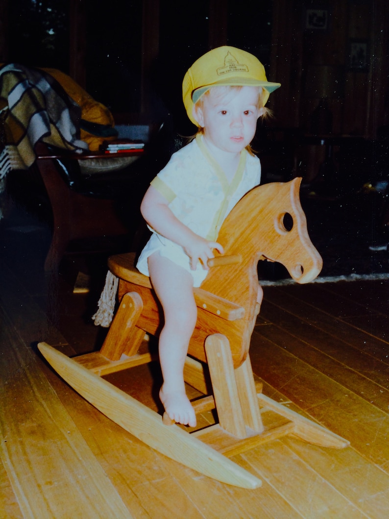 May include: A young child wearing a yellow and green baseball cap rides a wooden rocking horse. The horse is brown and has a white saddle. The child is wearing a white shirt with a yellow collar.