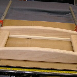 May include: Two red clamps are holding together a wooden frame. The frame is made of light wood and has a curved top and bottom. The frame is sitting on a brown surface.
