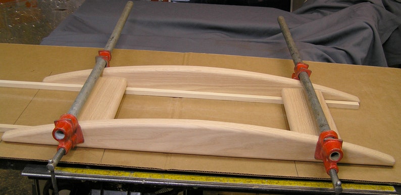 May include: Two red clamps are holding together a wooden frame. The frame is made of light wood and has a curved top and bottom. The frame is sitting on a brown surface.