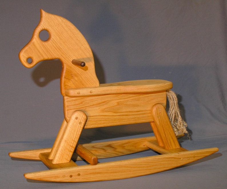 May include: A wooden rocking horse with a natural wood finish. The horse has a rope tail and a wooden saddle. The rocking base is made of wood and has a natural wood finish.