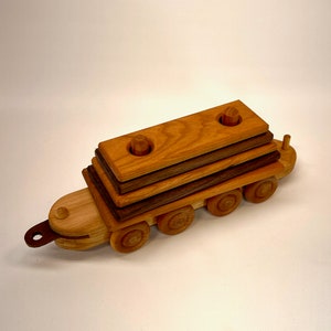 May include: A wooden toy train with a stack of four wooden logs on top. The train has six wheels and is made of light and dark wood.
