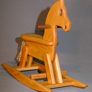 May include: A wooden rocking horse with a natural wood finish. The horse has a saddle and a rope mane and tail. The rocking base is made of wood and has a natural finish.