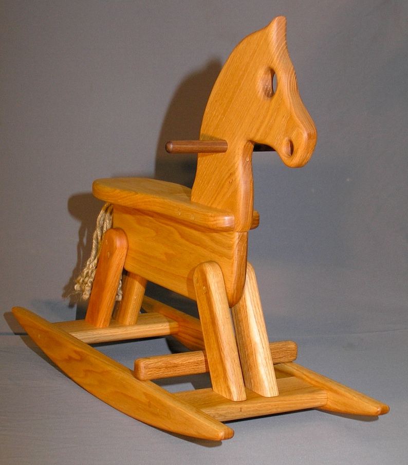 May include: A wooden rocking horse with a natural wood finish. The horse has a saddle and a rope mane and tail. The rocking base is made of wood and has a natural finish.