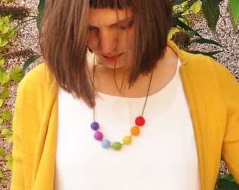 Puede incluir: Un collar colorido con bolas de fieltro en colores del arcoíris: morado, azul, verde, amarillo, naranja y rojo. El collar se lleva sobre una camiseta blanca y un cárdigan amarillo.