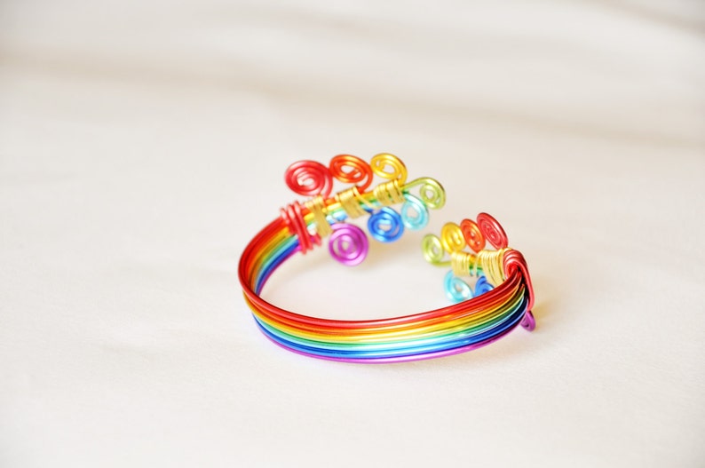May include: A rainbow-coloured wire bracelet with a decorative swirl design. The bracelet is made of multiple strands of wire, each a different colour of the rainbow.