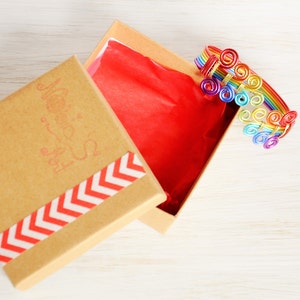 May include: A rainbow-coloured wire bracelet with a swirl design is displayed in a brown gift box with a red tissue paper lining and a white and red chevron pattern on the lid. The box lid has the words "Made with Love" printed on it.