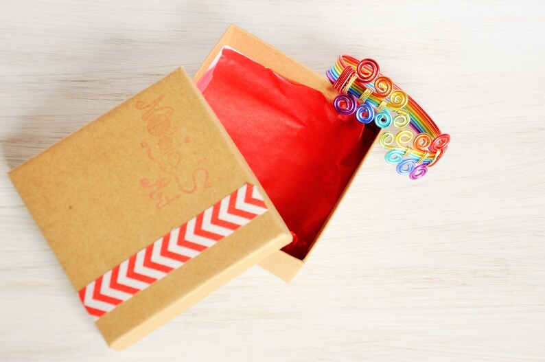 May include: A rainbow-coloured wire bracelet with a swirl design is displayed in a brown gift box with a red tissue paper lining and a white and red chevron pattern on the lid. The box lid has the words "Made with Love" printed on it.