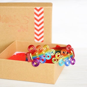 May include: A rainbow wire bracelet with a gold accent, in a brown box with a red interior and a white chevron pattern on the lid. The box has the text "Norie's Art" on the lid.