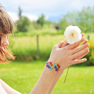 May include: A woman with long brown hair is holding a white flower in her hands. She is wearing a rainbow bracelet on her wrist.