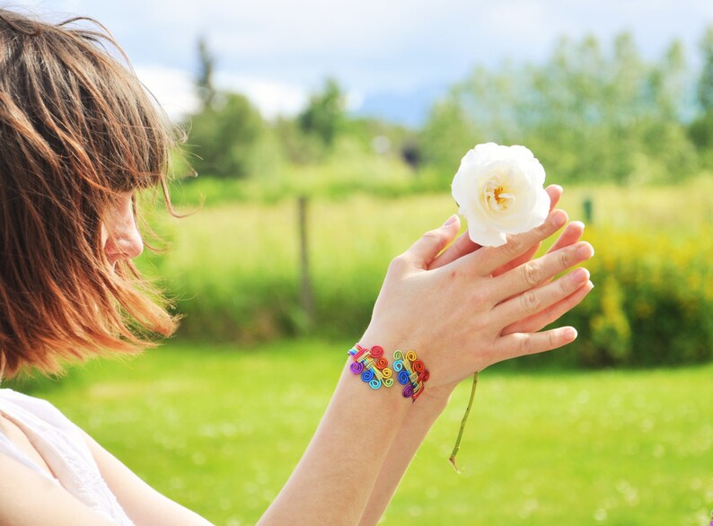 May include: A woman with long brown hair is holding a white flower in her hands. She is wearing a rainbow bracelet on her wrist.