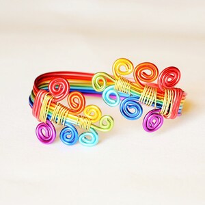 May include: A rainbow-coloured wire bracelet with a design of multiple swirls. The bracelet is made of thin wire and is a vibrant and colourful accessory.