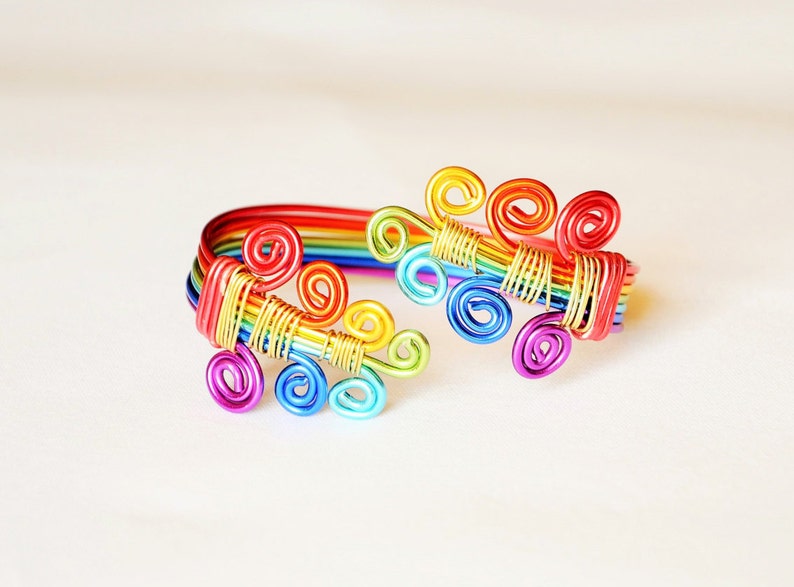 May include: A rainbow-coloured wire bracelet with a design of multiple swirls. The bracelet is made of thin wire and is a vibrant and colourful accessory.