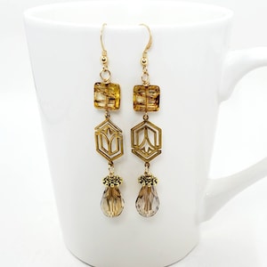 May include: A pair of gold-toned dangle earrings. Each earring features a square amber-colored bead, a geometric brass-colored pendant, and a teardrop-shaped crystal. The earrings are suspended from gold-colored ear wires.