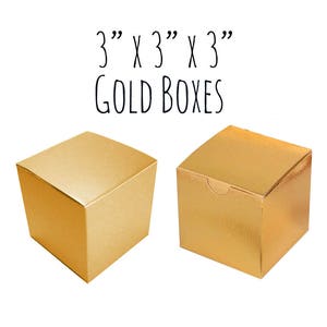 May include: Two gold 3" x 3" x 3" cardboard boxes. The boxes are empty and have a shiny finish.