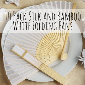 Op de afbeelding: Een set van 10 witte zijden en bamboe vouwvaandels. De vaandels liggen op een wit bord met een bruine achtergrond. De vaandels zijn gesloten en hebben een delicate, elegante uitstraling.