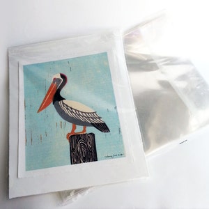 May include: A colourful illustration of a pelican perched on a wooden post against a light blue background. The pelican has a long orange beak, black and white feathers, and grey legs. The illustration is signed "Cathy Cox 2019".