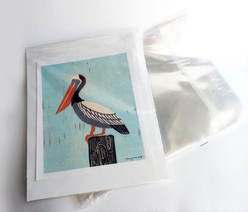 May include: A colourful illustration of a pelican perched on a wooden post against a light blue background. The pelican has a long orange beak, black and white feathers, and grey legs. The illustration is signed "Cathy Cox 2019".