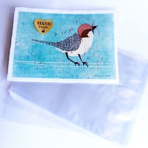 Puede incluir: Una impresi&oacute;n de arte hecha a mano con un p&aacute;jaro colorido con cabeza roja y cuerpo con estampado en blanco y negro. La impresi&oacute;n est&aacute; sobre un fondo azul con un borde blanco y un coraz&oacute;n dorado con el texto "HAND made" en &eacute;l.