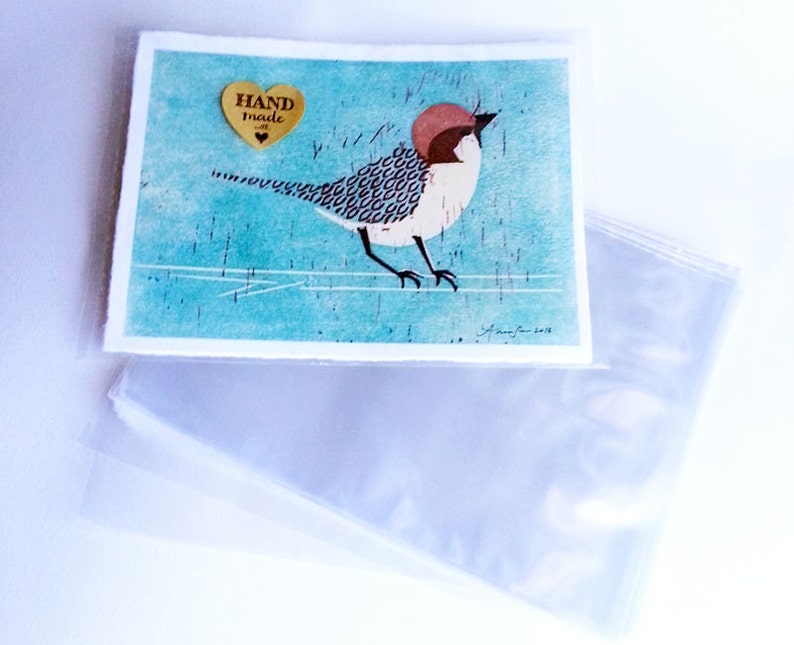 Puede incluir: Una impresi&oacute;n de arte hecha a mano con un p&aacute;jaro colorido con cabeza roja y cuerpo con estampado en blanco y negro. La impresi&oacute;n est&aacute; sobre un fondo azul con un borde blanco y un coraz&oacute;n dorado con el texto "HAND made" en &eacute;l.
