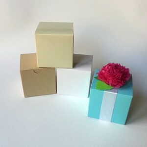 May include: Four small gift boxes in different colors. The boxes are stacked on top of each other. The top box is light blue and has a pink paper flower on top. The box below it is white. The box below that is brown. The bottom box is cream.