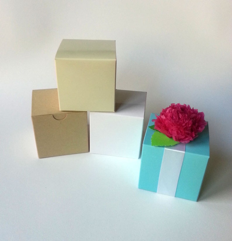 May include: Four small gift boxes in different colors. The boxes are stacked on top of each other. The top box is light blue and has a pink paper flower on top. The box below it is white. The box below that is brown. The bottom box is cream.