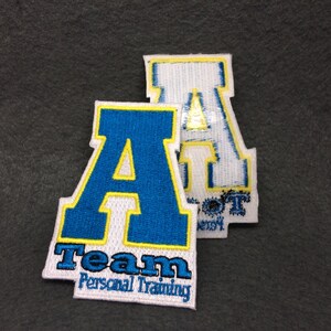 May include: Two white felt patches with blue and yellow embroidered letters. The larger patch says "Team Personal Training" and features a large blue "A" with a yellow outline. The smaller patch features a white "A" with a yellow outline.