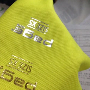 May include: Close-up of a yellow fabric with a silver and gold "Ped" logo. The logo is repeated twice, once in silver and once in gold. The text "Size SX" is also visible on the fabric.