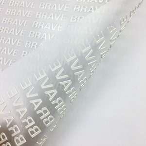 以下が含まれることがあります： 白い半透明シートのクローズアップ。シートには「BRAVE」という単語が繰り返しエンボス加工されています。