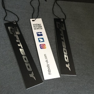 Könnte beinhalten: Drei schwarze und weiße Hängeetiketten mit dem Text "Fitbody" in silberner Schrift. Das mittlere Etikett hat einen QR-Code, Facebook-, Twitter- und Instagram-Symbole sowie den Text "Fitbody-uk.com".