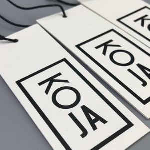 May include: Three white rectangular hang tags with black string attached. Each tag has a black square with the text "KOJA" in black lettering.