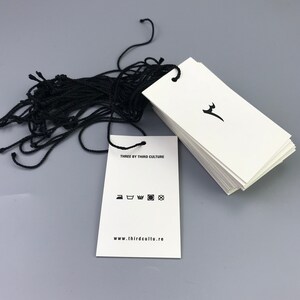 May include: A stack of white hang tags with black string attached. The tag has the text "THREE BY THIRD CULTURE" and a website address. The tag also has a black and white graphic of a stylized deer head and a set of care instructions.