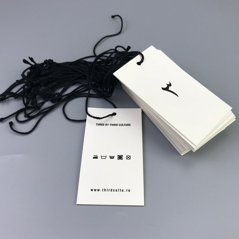 May include: A stack of white hang tags with black string attached. The tag has the text "THREE BY THIRD CULTURE" and a website address. The tag also has a black and white graphic of a stylized deer head and a set of care instructions.