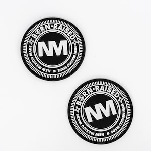 May include: Two black round stickers with white text. The text reads "BORN-RAISED" around the top, "NM" in the center, and "NEW MEXICO MADE" around the bottom. The stickers have a white border and are on a white background.