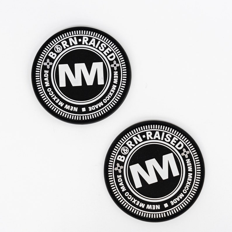 May include: Two black round stickers with white text. The text reads "BORN-RAISED" around the top, "NM" in the center, and "NEW MEXICO MADE" around the bottom. The stickers have a white border and are on a white background.