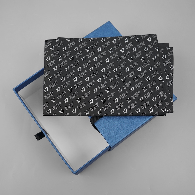 May include: A blue gift box with a black and white patterned tissue paper insert. The tissue paper has the words "Be More Kinetic" repeated in a pattern.