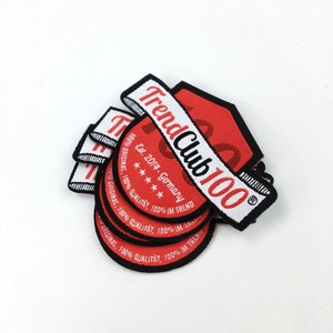 May include: A stack of red and white woven patches with black trim. The patches feature the text "Trend Club 100" and "Est. 2014 - Germany".