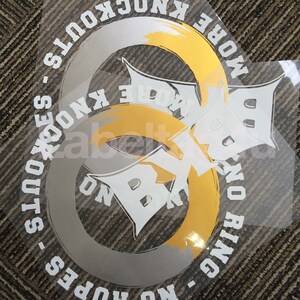 Op de afbeelding: Een witte en gouden vinyl sticker met de tekst "More Knockouts - No Ring - No Hopes" en "More Knockouts" in een cirkel. De sticker heeft een gestileerde afbeelding van een oneindigheidssymbool.