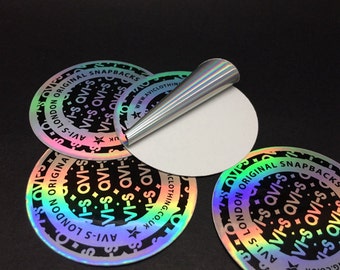 Può includere: Quattro adesivi rotondi olografici iridescenti con testo nero che recita "AVI-S LONDON ORIGINAL SNAPBACKS" e "AVI-S QVI-S" ripetuto attorno al cerchio. Gli adesivi hanno un retro bianco e una stella con il testo "CLOTHING.CO.UK" e "WWW.AVICLOTHING.CO.UK" stampato sotto la stella.