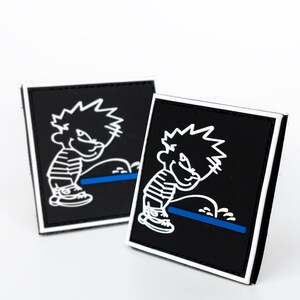 May include: Two square black patches with a white border. Each patch features a cartoon illustration of a boy urinating, with a blue line and white accents. The boy is wearing a striped shirt. The patches are displayed on a white background.