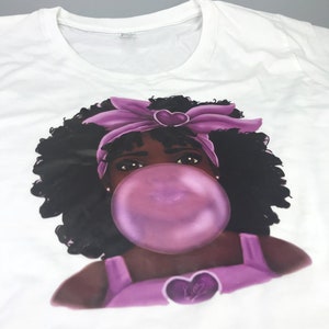 May include: White t-shirt with a graphic of a young girl with dark curly hair wearing a purple headband and blowing a large bubblegum bubble. The bubblegum is purple and the girl has brown eyes. The graphic also includes a purple heart with the word "Love" inside.
