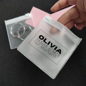 Può includere: Sacchetti trasparenti per gioielli in plastica con chiusura a zip. Una borsa contiene orecchini a cerchio argentati. L'altra borsa ha il testo "OLIVIA DESIGN STUDIO" stampato in nero. Le borse sono tenute in mano.