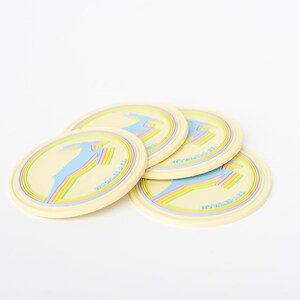 May include: Four cream-colored round coasters with a colorful design. Each coaster features a blue figure with a rainbow accent and the text "MOONRISE CO." in a circular border. The coasters are stacked slightly.