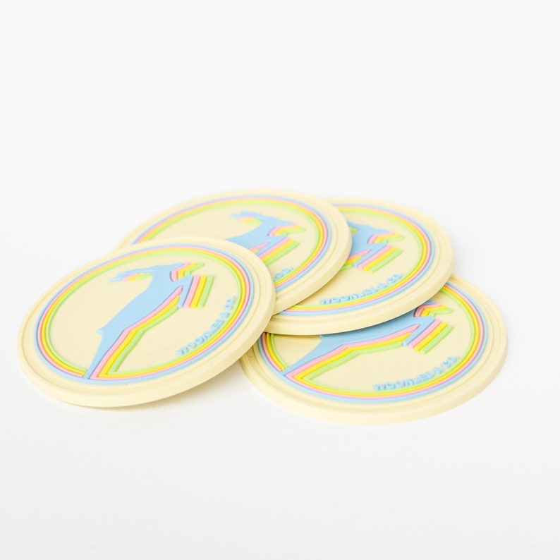 May include: Four cream-colored round coasters with a colorful design. Each coaster features a blue figure with a rainbow accent and the text "MOONRISE CO." in a circular border. The coasters are stacked slightly.
