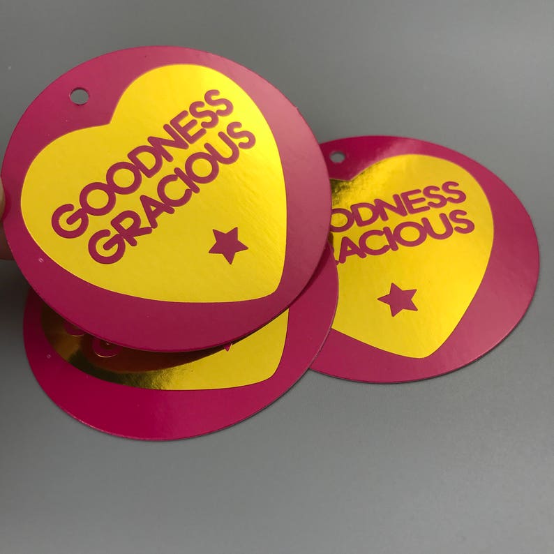Pu&ograve; includere: Tre etichette regalo rotonde rosa con cuori in foglia d'oro. I cuori hanno il testo "GOODNESS GRACIOUS" in lettere rosa. Ogni etichetta ha una stella dorata al centro del cuore.