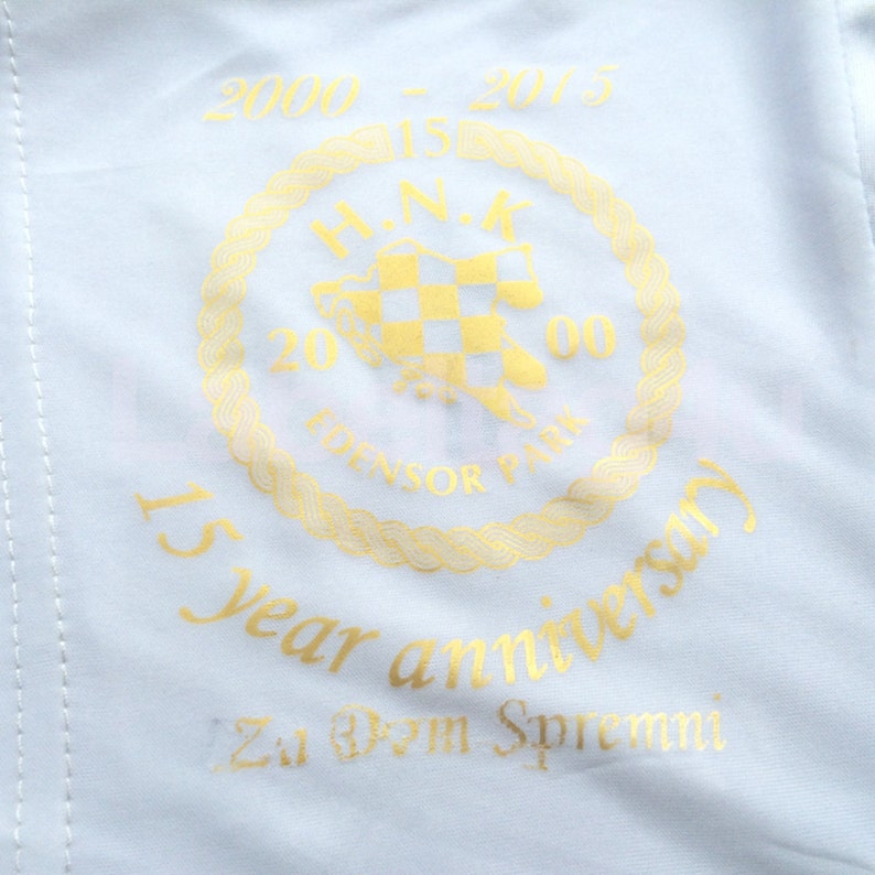 Op de afbeelding: Een witte stoffen patch met gouden letters die "2000 - 2015 15 year anniversary Zu Dem Spremni" luidt en een gouden cirkelvormig logo met de tekst "H.N.K. 20 EDENSOR PARK 00".
