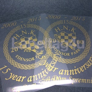 Op de afbeelding: Twee gouden stickers met een zwart-wit geruit ontwerp in een cirkel met de tekst "H.N.K. 2000 - 2015 EDENSOR PARK" en de tekst "15 year anniversary" eronder.