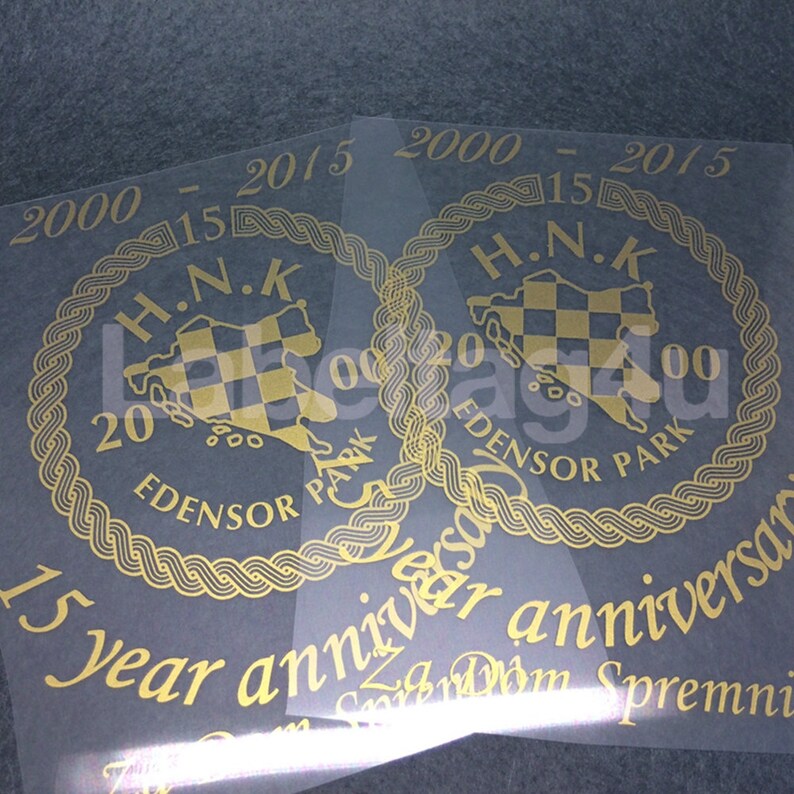 Op de afbeelding: Twee gouden stickers met een zwart-wit geruit ontwerp in een cirkel met de tekst "H.N.K. 2000 - 2015 EDENSOR PARK" en de tekst "15 year anniversary" eronder.