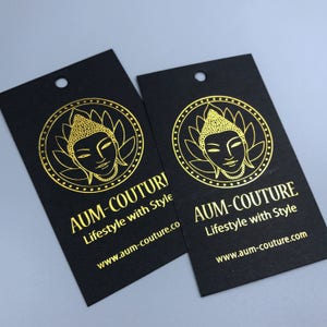 Pu&ograve; includere: Due etichette regalo nere con lettering dorato e un'immagine dorata di una testa di Buddha. Il testo sulle etichette recita "AUM-COUTURE Lifestyle with Style" e "www.aum-couture.co".
